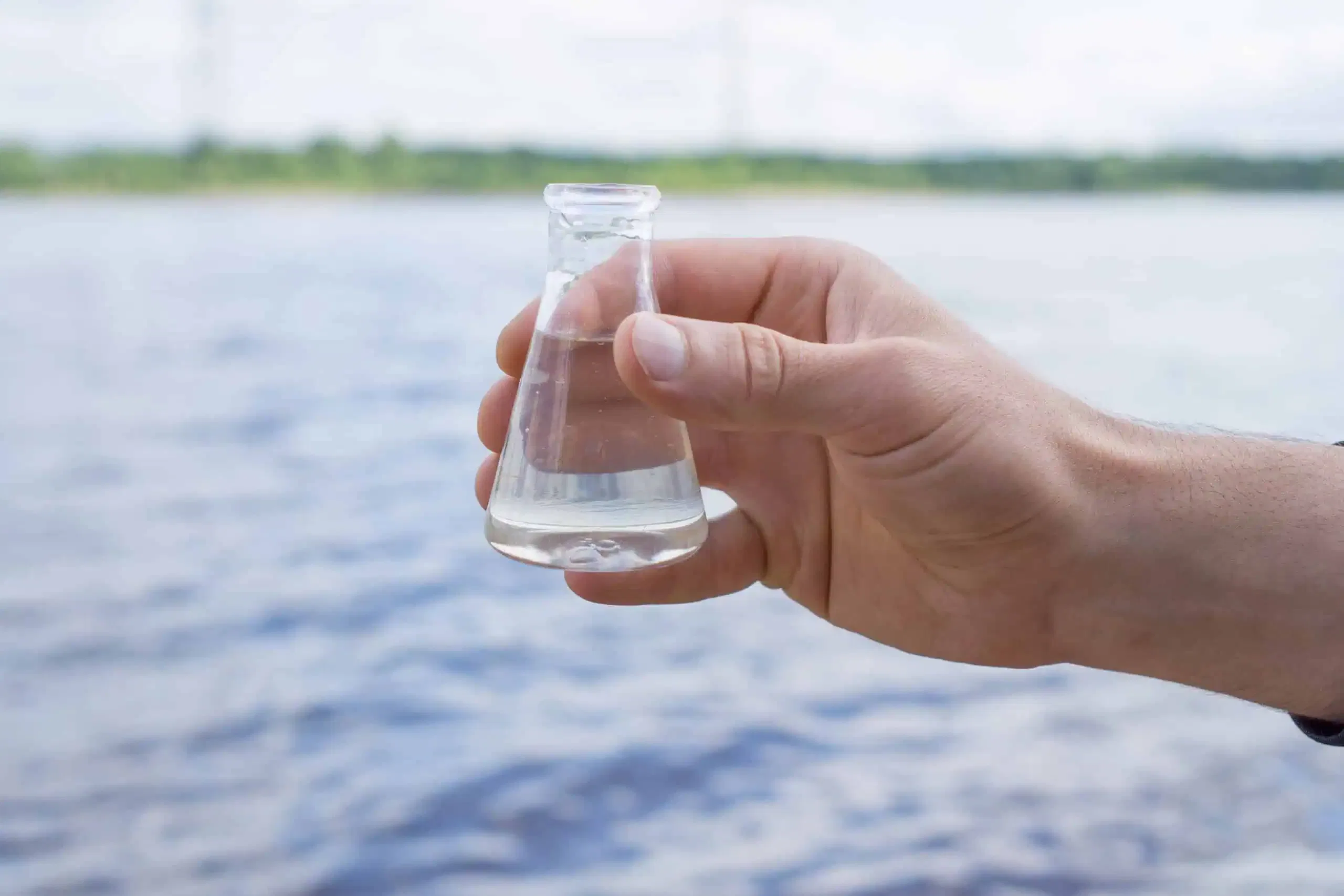 A hand holding a small glass flask with clear liquid in front of a pristine body of water showcases the epitome of quality.