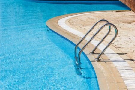 A stainless steel pool ladder descends into a clear blue swimming pool, bordered by a stone deck—a pristine example of quality and maintenance offered through our Commercial Pool Services.