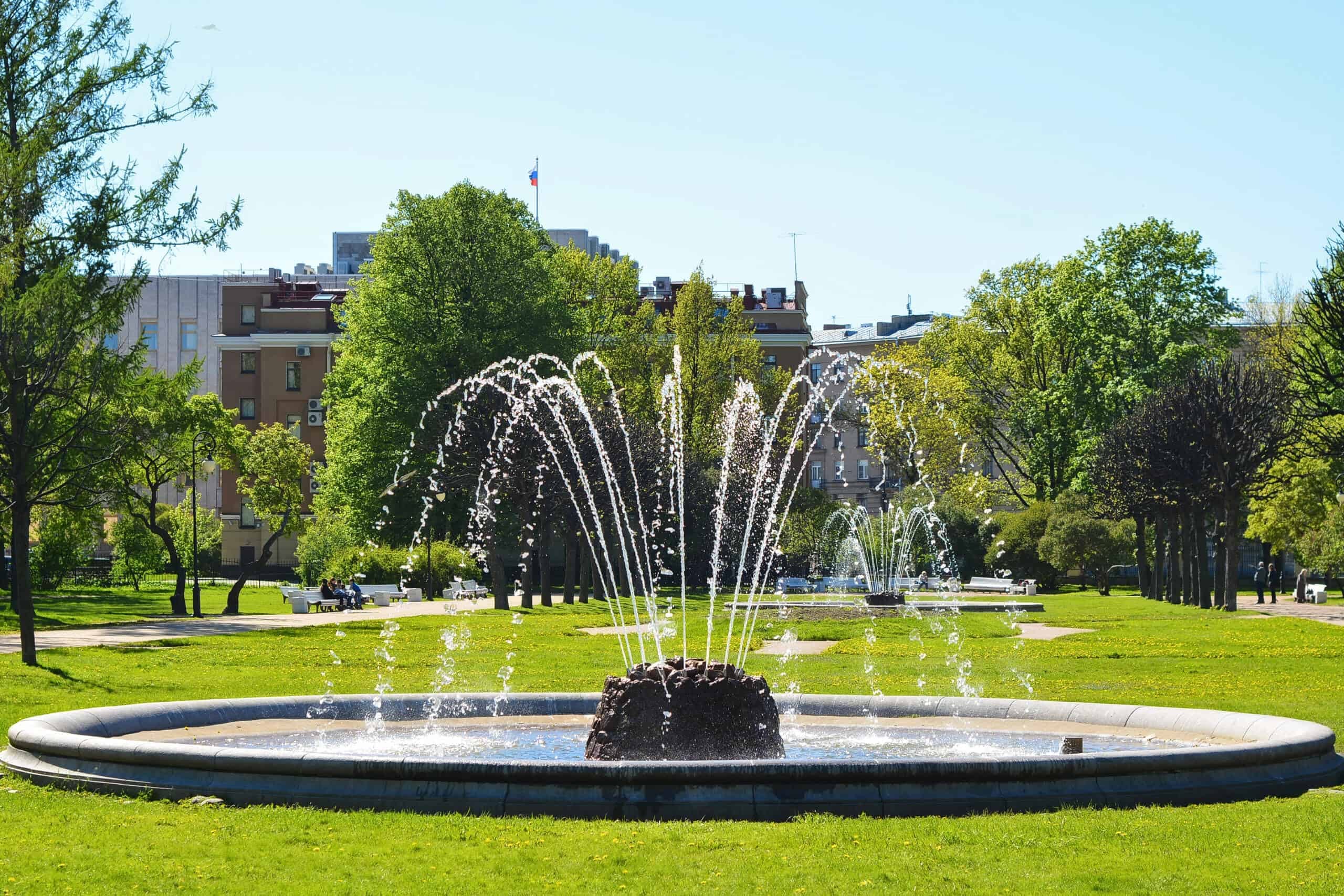 A circular fountain in a park with water streams spraying upwards, surrounded by green grass, trees, and buildings in the background under a clear blue sky—exemplifying expert fountain services.