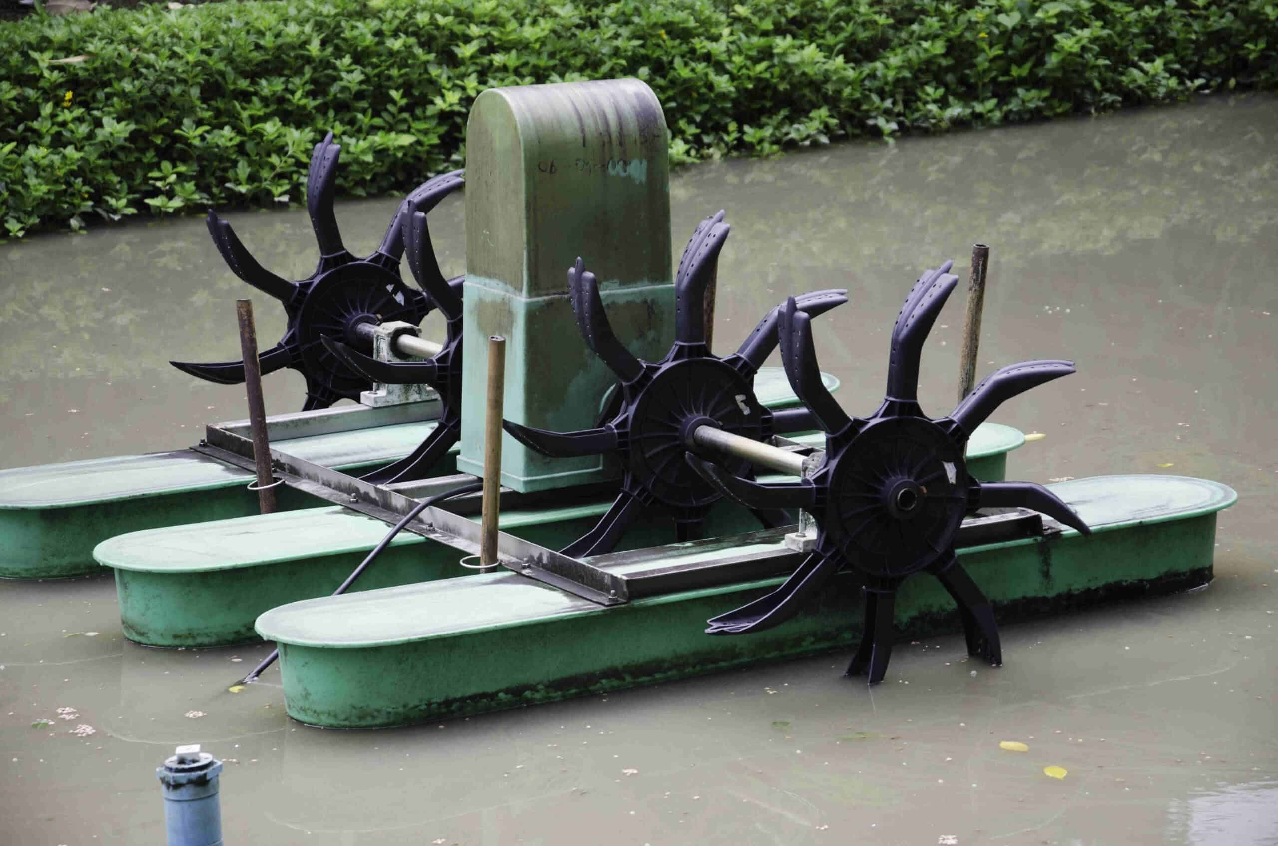 A close-up of a green paddle wheel aerator in a pond, featuring black rotating paddles and a central motor housing, with greenery in the background. This lake aeration device ensures optimal water quality and oxygen levels for aquatic life.