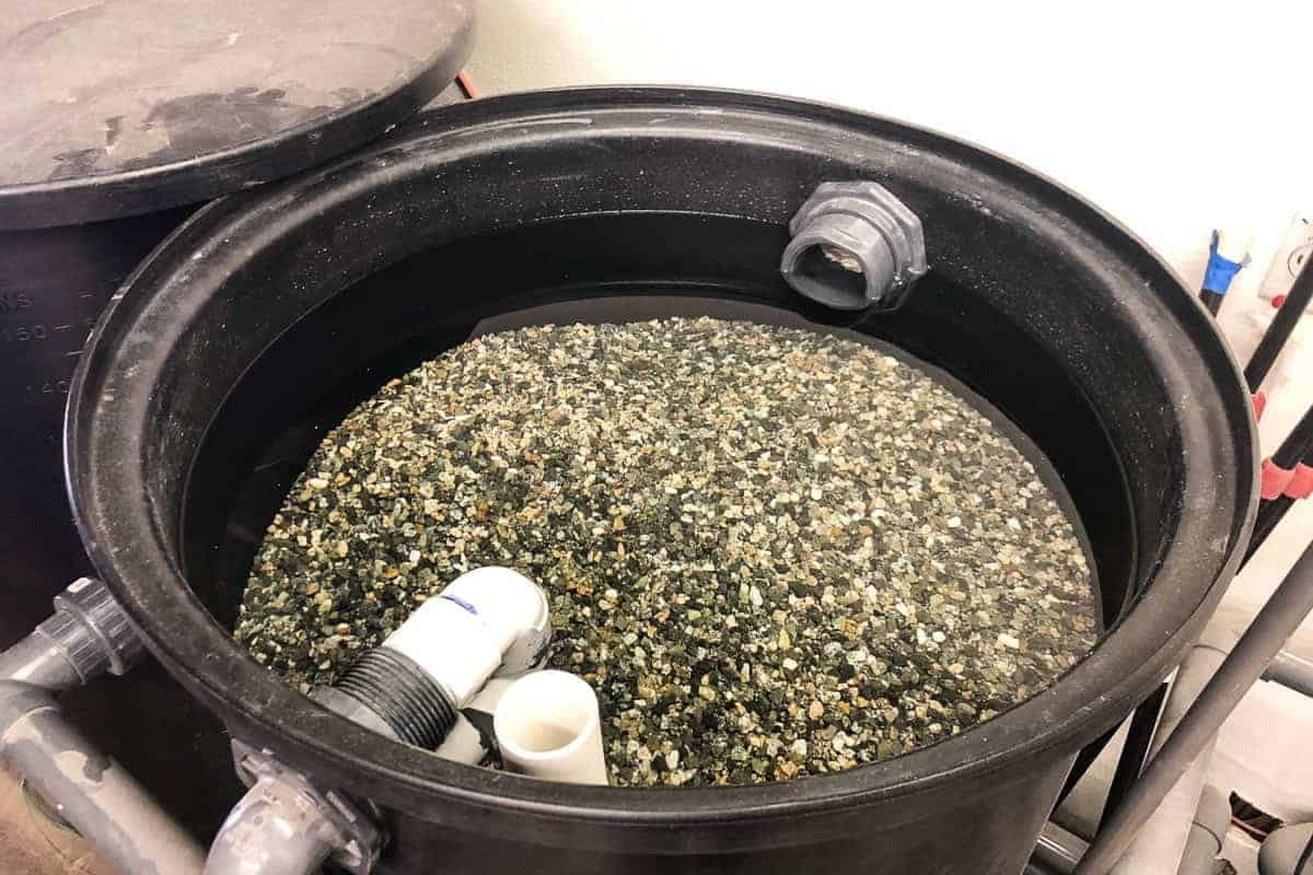 A large black cylindrical container is filled with a mixture of assorted gravel and water, featuring several PVC piping components.