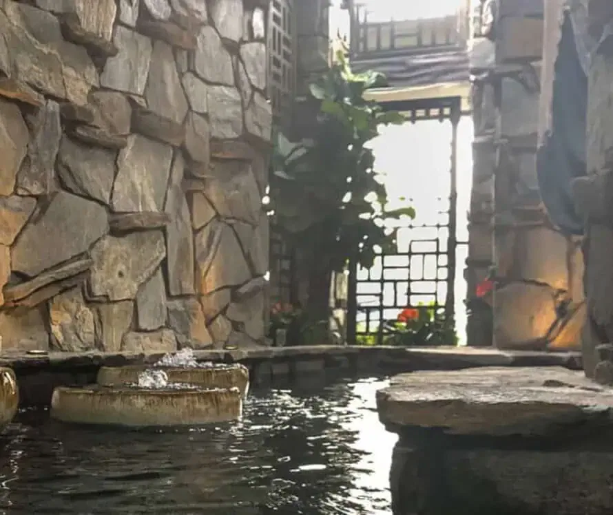 A serene stone statue stands by a shaded indoor water feature, surrounded by stone walls and greenery, with sunlight filtering through in the background.