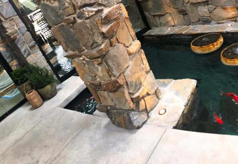 Indoor pond with koi fish and a stone statue. The background includes a stone wall and pillars, and the pond features three round water features.