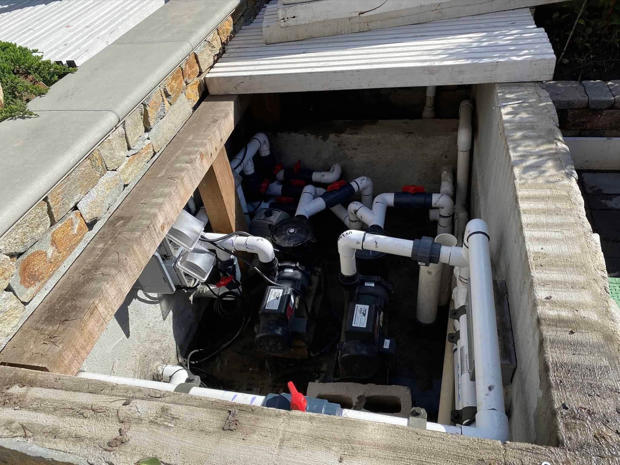 A maintenance access pit with various plumbing pipes, valves, and two black pump units inside. Some pipes are marked with red handles, and wiring is also visible; this setup is integral to the project's overall infrastructure.
