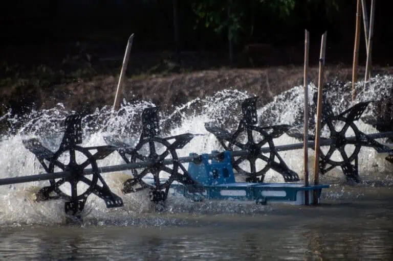 Water wheels rotating in a pond, generating splashes. The wheels, which appear to be part of pond maintenance equipment, are connected to a metal structure likely for mechanical or agricultural purposes. In the background, plants are partially visible.