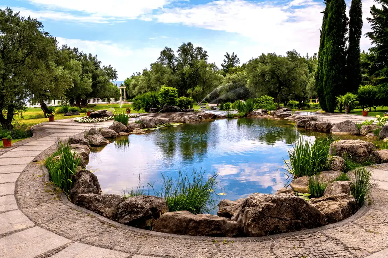 A landscaped garden pond surrounded by large rocks, green plants, and trees, with a stone walkway circling the water under a partly cloudy sky—perfect inspiration for your next Blog Post Template.