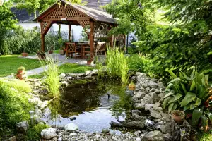 A small California garden pond with rocks and tall plants is in the foreground, while a wooden gazebo and outdoor furniture surrounded by greenery await in the background—perfect for relaxing or trying DIY algae control.