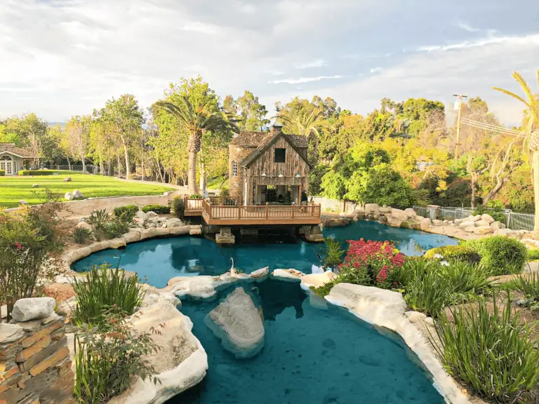 A rustic wooden house sits beside a large, landscaped pond with rocks and plants in a lush garden setting—ideal for enjoying nature if you take steps to prevent fish death and keep the clear blue water healthy for all pond life.