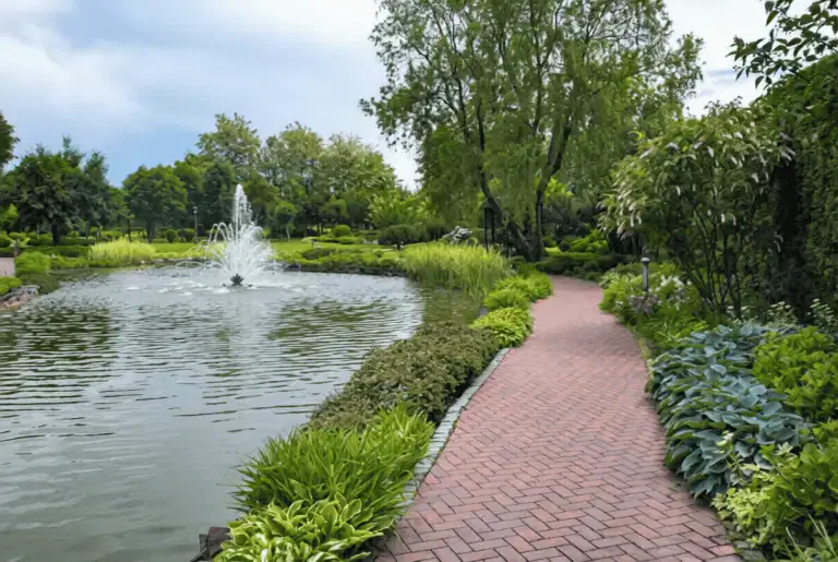 A brick path curves beside a pond with a fountain, surrounded by lush green plants and neatly landscaped gardens—an idyllic spot enhanced by organic pond algae control for optimal California pond maintenance under a partly cloudy sky.