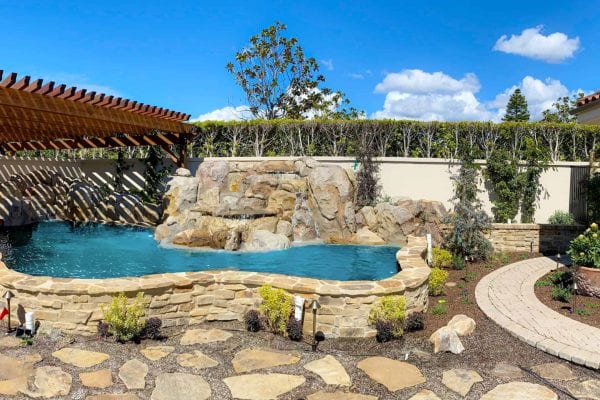 A backyard with a small stone swimming pool, a rock waterfall feature, and a wooden pergola makes for an exquisite project. A curved stone walkway and various plants decorate the area.