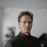 A man with short brown hair and dressed in a dark suit stands indoors, looking directly at the camera. The background is slightly blurred, showing some indistinct black-and-white photographs; it almost feels like a quintessential blog post title image.