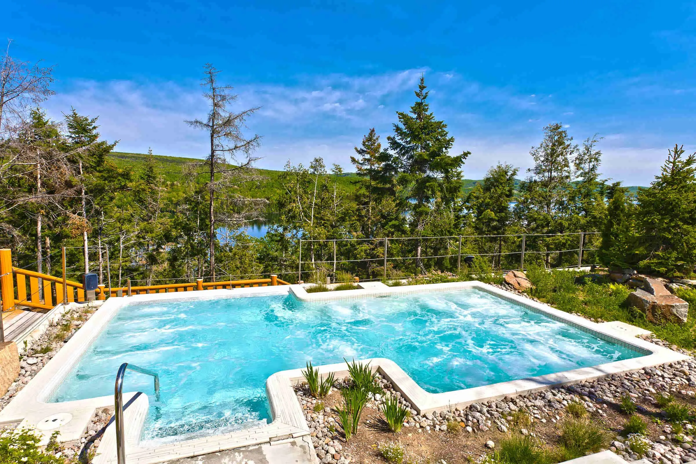Outdoor pool with clear blue water, surrounded by trees and a view of a distant lake under a bright blue sky. A metal handrail is visible on the left side of the pool, enhancing the serene water feature at our luxurious hotel.