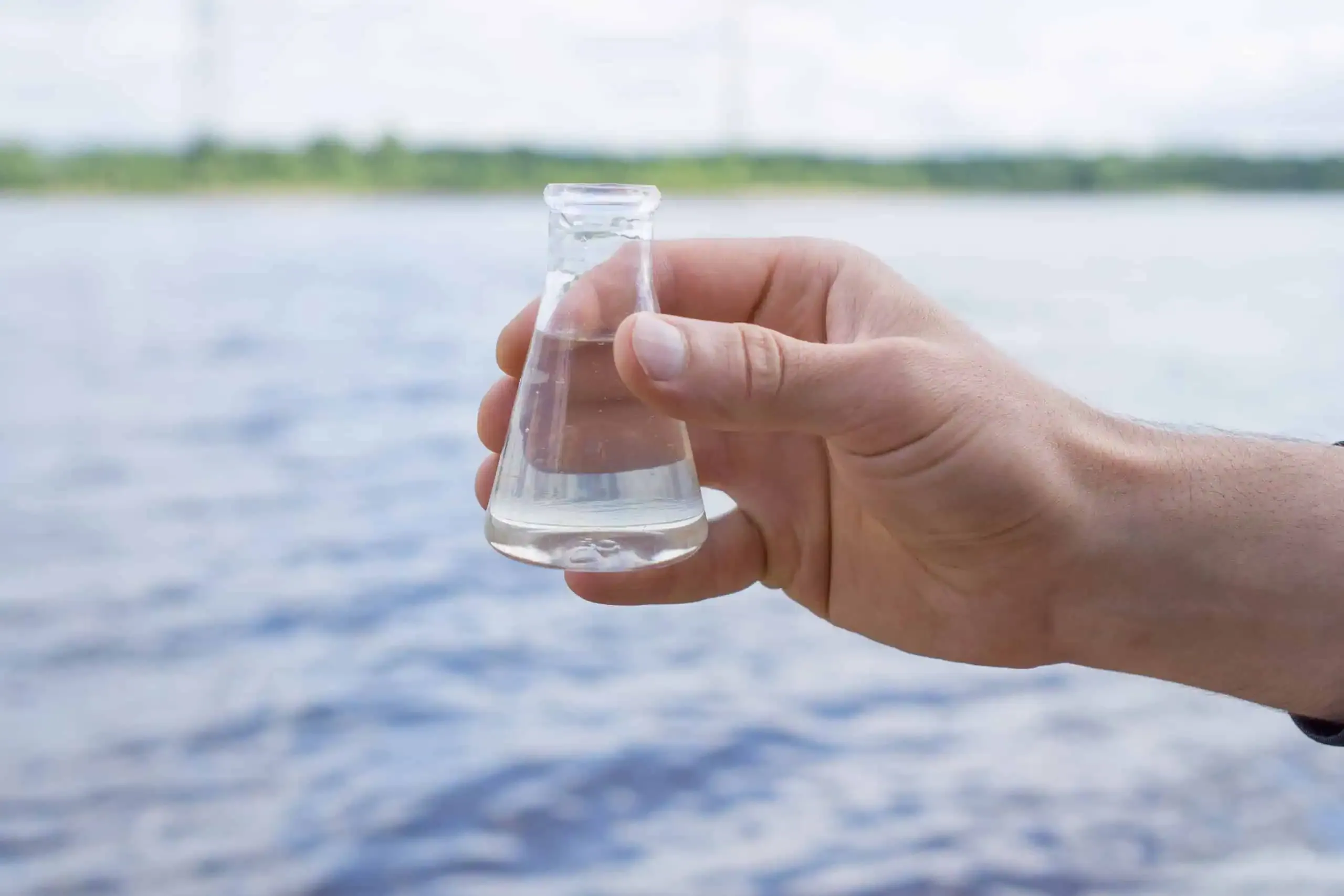A hand holding a small glass flask with clear liquid in front of a pristine body of water showcases the epitome of quality.