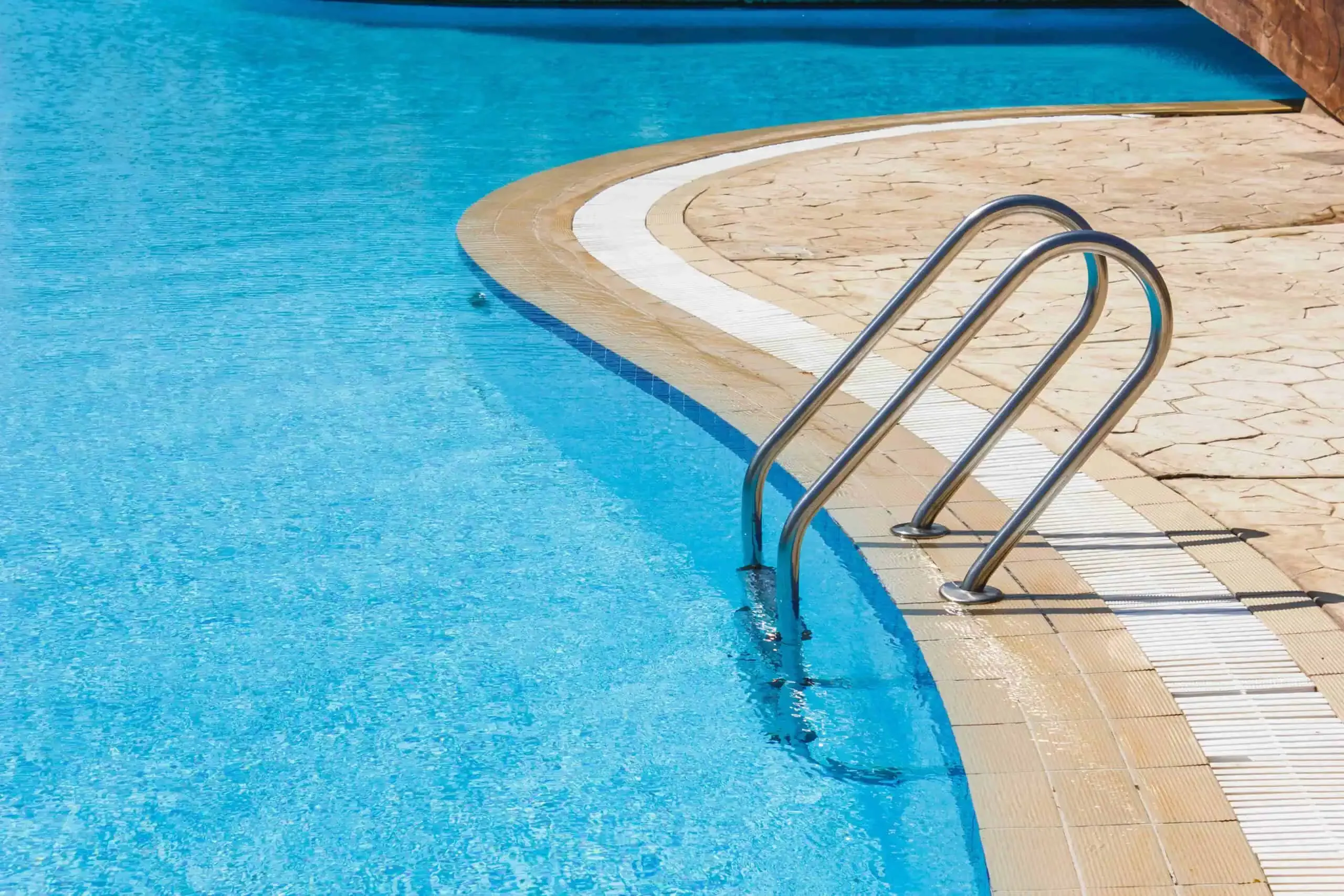 A stainless steel pool ladder descends into a clear blue swimming pool, bordered by a stone deck—a pristine example of quality and maintenance offered through our Commercial Pool Services.