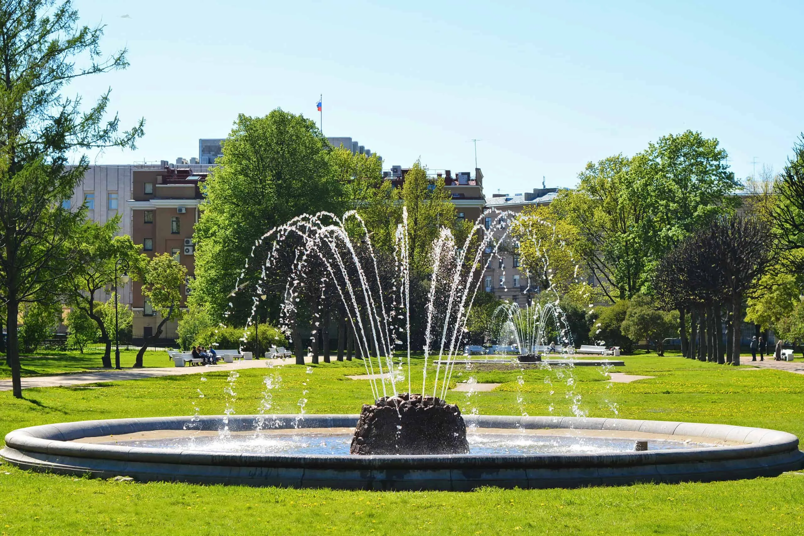 A circular fountain in a park with water streams spraying upwards, surrounded by green grass, trees, and buildings in the background under a clear blue sky—exemplifying expert fountain services.