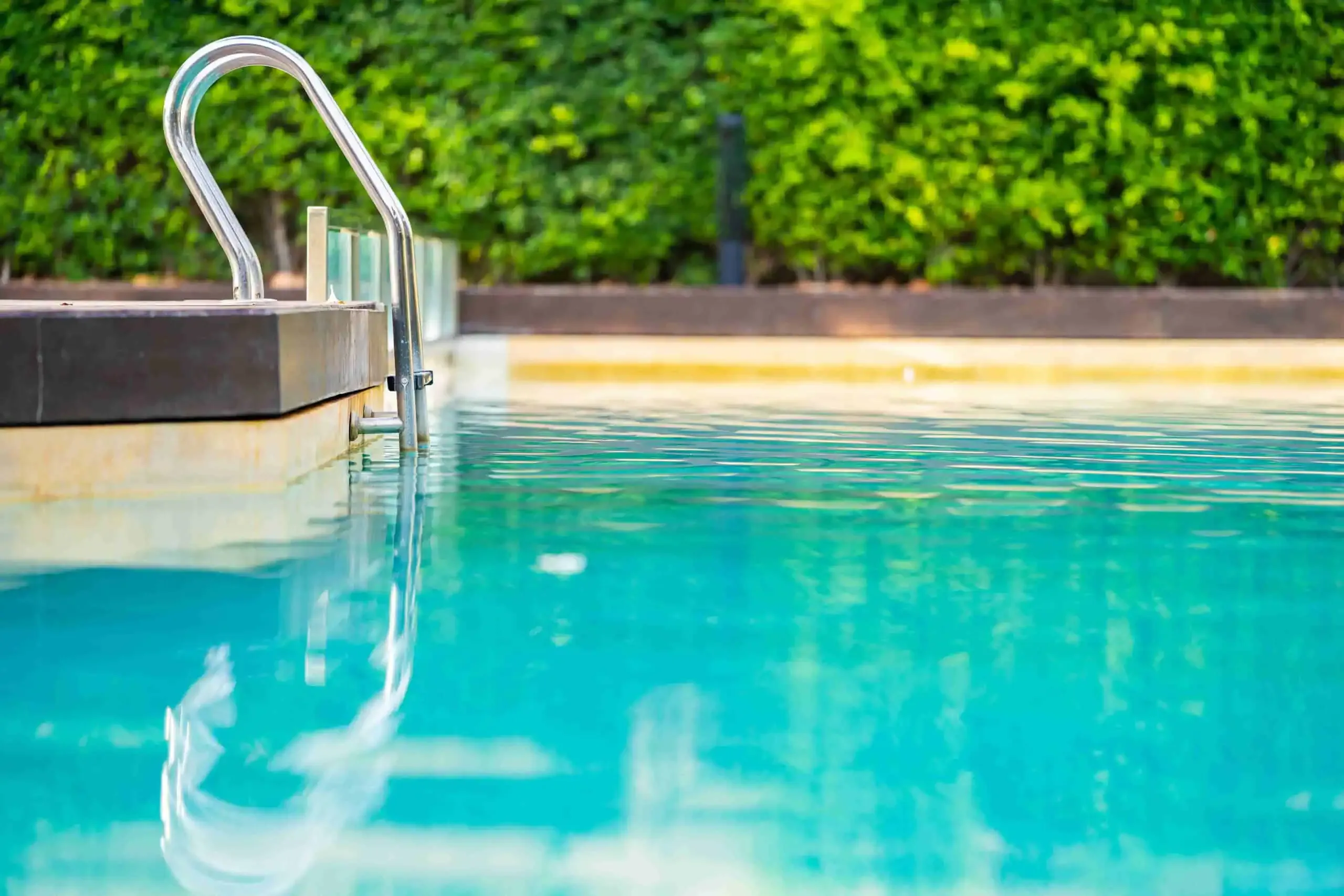 A clear swimming pool with metal handrails and a clean, green shrub border in the background, thanks to professional pool care.