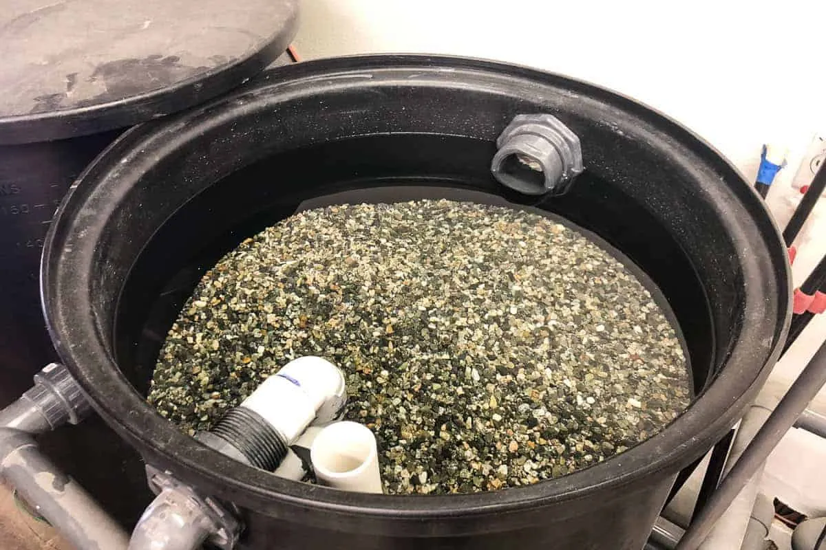 A large black cylindrical container is filled with a mixture of assorted gravel and water, featuring several PVC piping components.