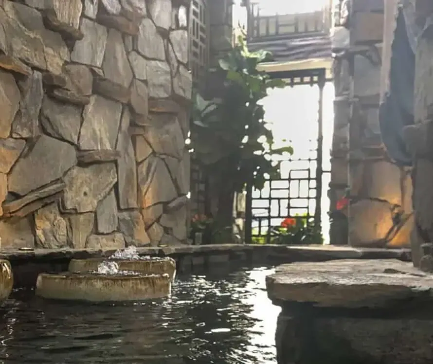 A serene stone statue stands by a shaded indoor water feature, surrounded by stone walls and greenery, with sunlight filtering through in the background.