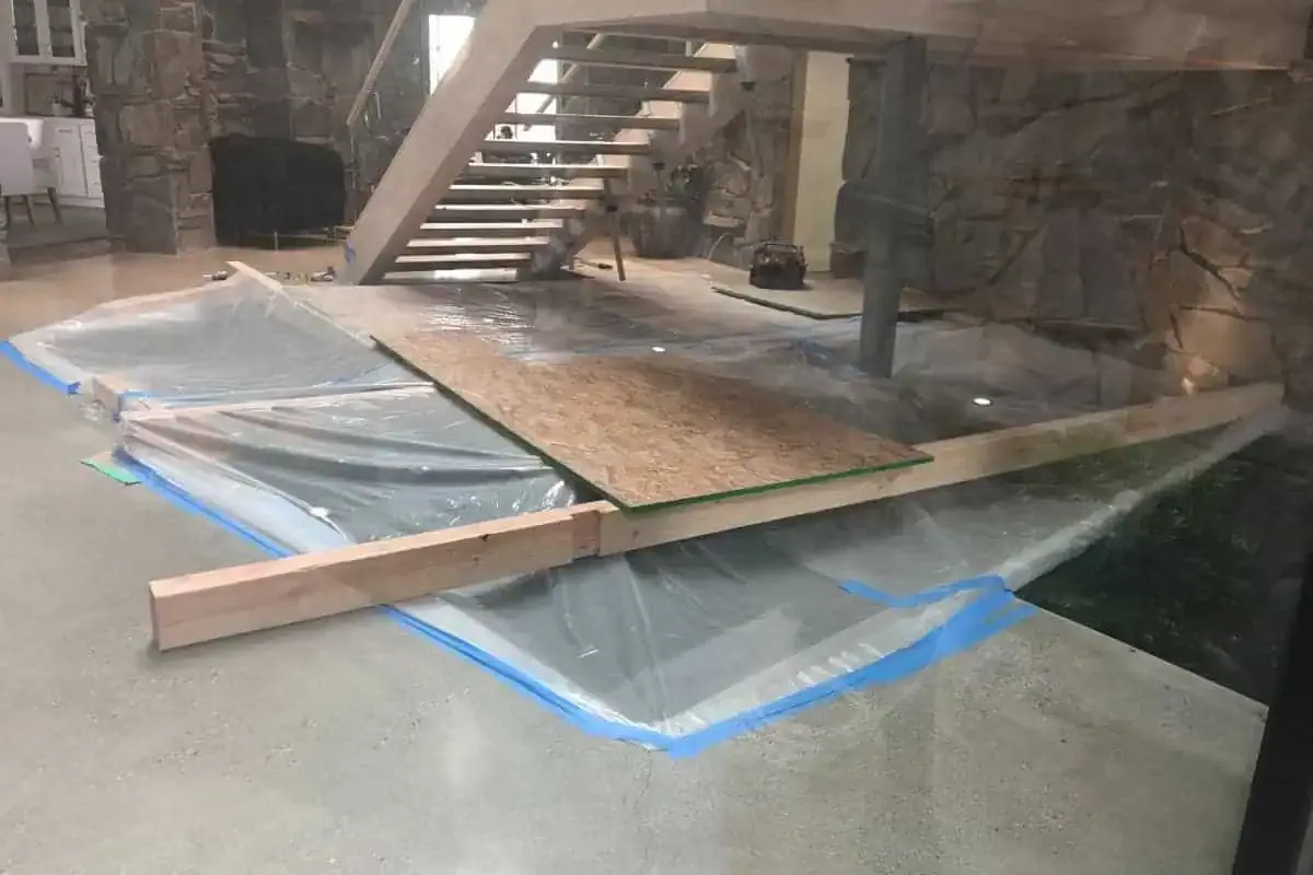 A work area is set up under a staircase with plastic sheets, wooden planks, and tools suggestive of a renovation or construction project in progress.