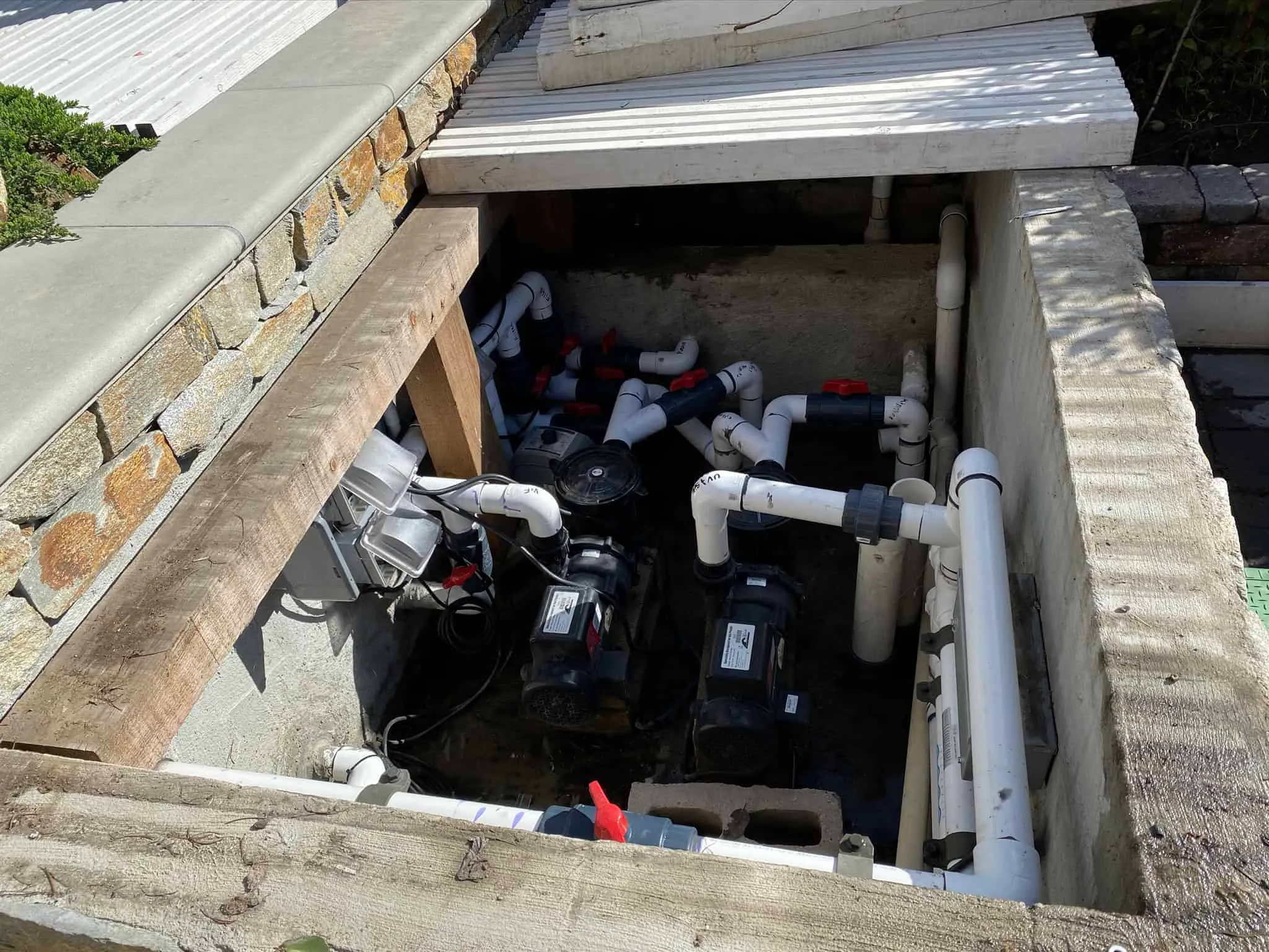 A maintenance access pit with various plumbing pipes, valves, and two black pump units inside. Some pipes are marked with red handles, and wiring is also visible; this setup is integral to the project's overall infrastructure.
