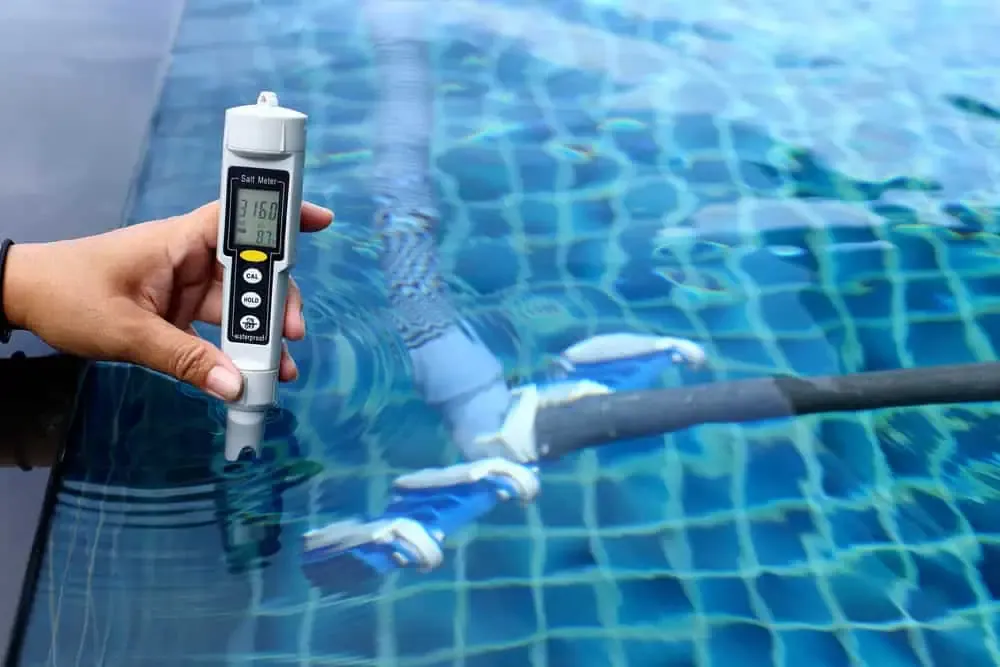 A hand holding a salt meter above the water in a swimming pool, checking for optimal conditions to get rid of pool algae. The meter's display reads 3166 ppm. A portion of a pool cleaning device is visible underwater.