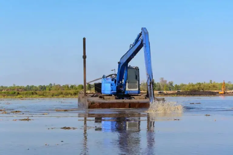 A blue excavator, essential for pond maintenance equipment, operates in shallow water with its arm extended and digging. The background includes distant foliage under a clear blue sky.