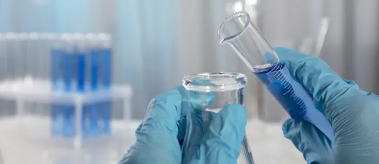 A person wearing blue gloves pours liquid from a small beaker into a larger one in a laboratory setting, demonstrating pond water quality testing, with test tubes containing blue liquid visible in the background.
