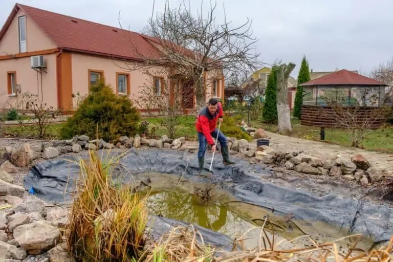 A person in a red jacket stands by a small California backyard pond, using a net to clean debris and manage pond algae, with a house and garden visible in the background.