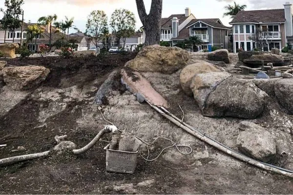 An empty pond with large boulders, pipes, aerators, empty filtration, and hoses. Residential houses are visible in the background as workers lay the groundwork for a new business center.